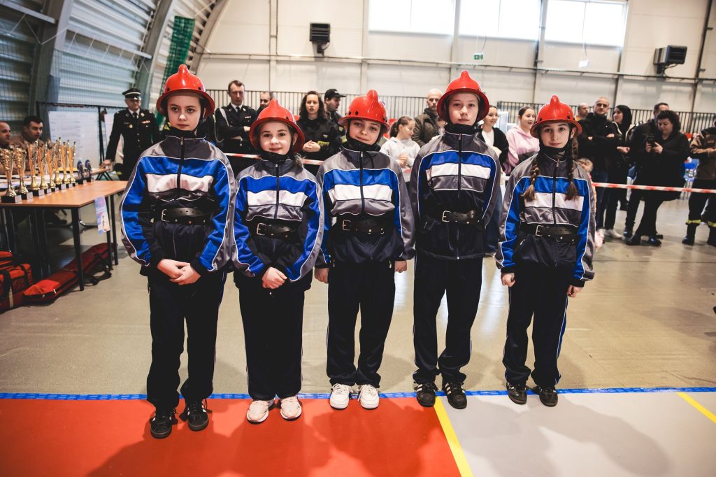 Pięć dziewcząt w czerwonych hełmach i granatowo-szaro-niebieskich mundurkach sportowych stoi w rzędzie na hali z widoczną publicznością i stołem z nagrodami w tle.