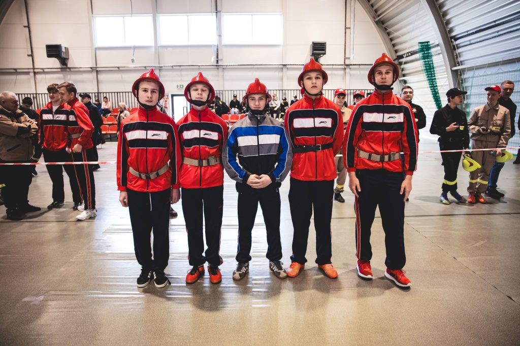 Pięciu nastolatków w czerwonych kurtkach i czerwonych hełmach stoi w rzędzie w hali sportowej, w tle ludzie i trybuny z czerwonymi siedzeniami.