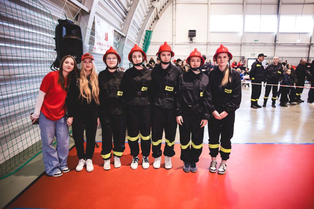 Siedem młodych kobiet, pięć w czarnych mundurach strażackich z czerwonymi hełmami, dwie w casualowych ubraniach, stoją na czerwonej podłodze w hali sportowej.