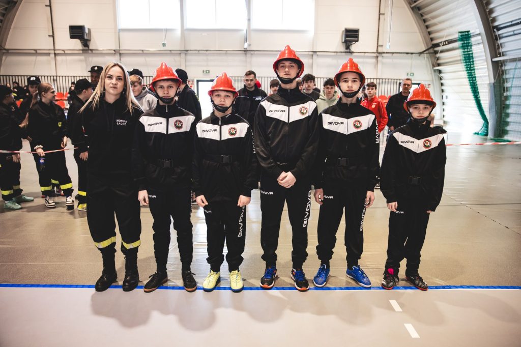 Grupa młodych strażaków w czarnych mundurach i czerwonych hełmach oraz kobieta w czarnym ubraniu stojąca w hali sportowej na jasnej podłodze.