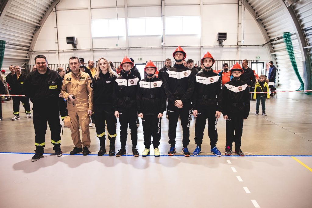Grupa ludzi w sali gimnastycznej, pięcioro dzieci w czarno-białych dresach i czerwonych hełmach, obok dorośli w strojach strażackich i cywilnych, jasne wnętrze hali.