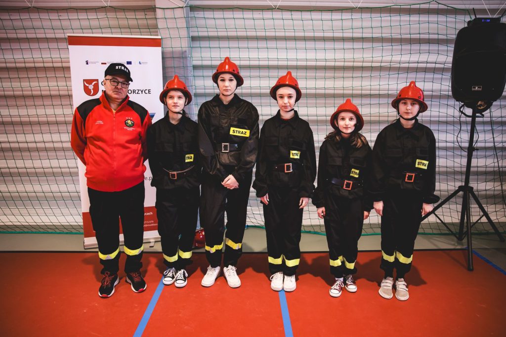 Pięć dzieci w czarnych uniformach z odblaskowymi paskami i czerwonych hełmach oraz dorosły mężczyzna w czerwonej bluzie i czapce, stoją na czerwonej podłodze przed zieloną siatką.