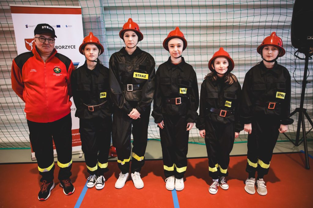 Grupa osób w czarnych mundurach z żółtymi naszywkami "STRAŻ" i czerwonych hełmach, obok mężczyzna w czerwonej kurtce i czapce z napisem "STRAŻ", na tle zielonej siatki.