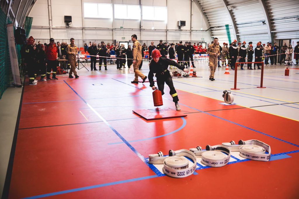 Młoda osoba w czarnym ubraniu strażackim i czerwonym hełmie biegnie po czerwonej hali sportowej z gaśnicą, w tle ludzie w mundurach i czerwonych hełmach obserwują zawody.