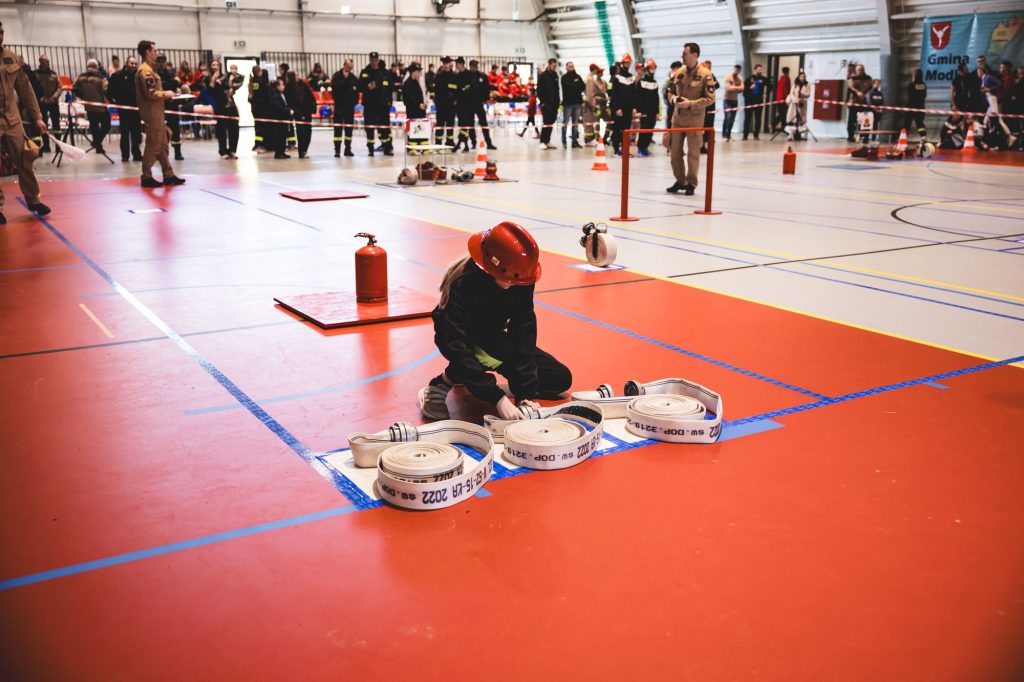 Osoba w czerwonym hełmie i ciemnym ubraniu zwija białe węże strażackie na pomarańczowej podłodze hali sportowej, w tle grupa ludzi i sprzęt strażacki.