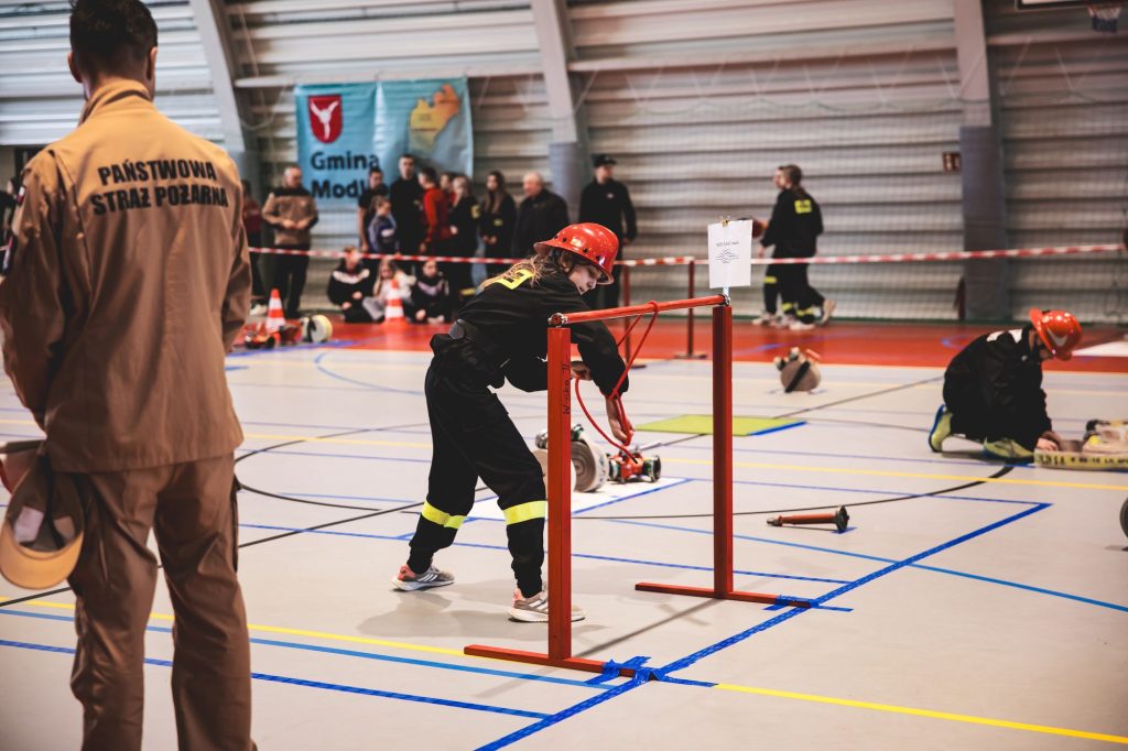 Dziecko w czarnym ubraniu i czerwonym hełmie manewruje liną na hali sportowej, obok stoi strażak w beżowym uniformie z napisem Państwowa Straż Pożarna.