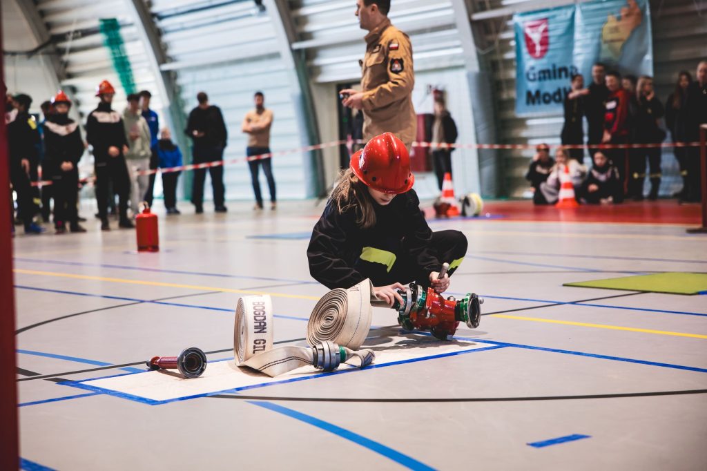 Dziewczynka w czerwonym hełmie i czarnym ubraniu z żółtymi paskami montuje element strażackiego węża rozłożonego na hali sportowej, w tle grupa ludzi.