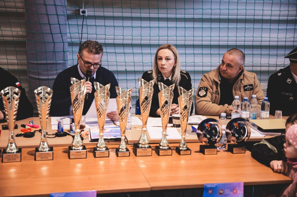 Stół z dziesięcioma pucharami i dwoma statuetkami, za nim siedzi pięć osób, w tym mężczyzna z mikrofonem i kobieta w mundurze, tło z zieloną siatką.