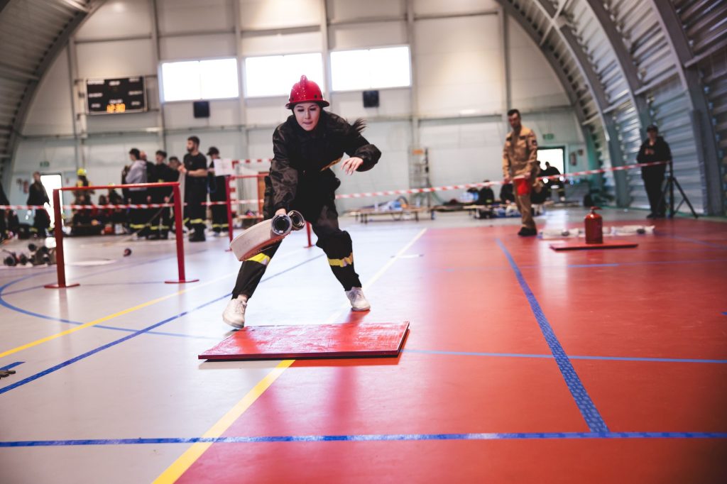 Kobieta w czerwonym hełmie i czarnym ubraniu trzyma rozwinięty wąż strażacki na hali sportowej z czerwoną i szarą podłogą, w tle inni ludzie.