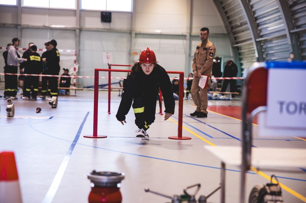 Młoda osoba w czarnym ubraniu i czerwonym hełmie przechodzi pod czerwoną poprzeczką na hali sportowej, w tle ludzie w mundurach i sprzęt strażacki.