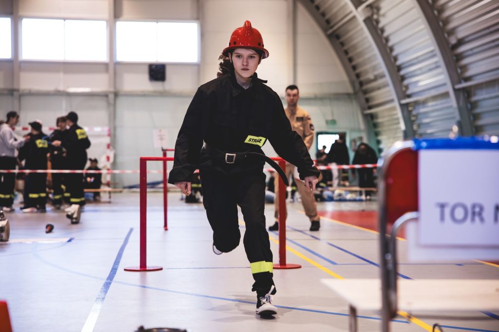 Młoda osoba w czarnym stroju strażaka i czerwonym hełmie biegnie w hali sportowej, w tle ludzie i metalowa konstrukcja.