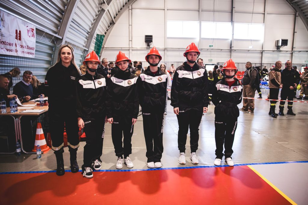 Pięć młodych osób w czarnych strojach i czerwonych hełmach oraz kobieta w czarnym ubraniu stoi na hali sportowej na czerwonym i szarym podłożu.