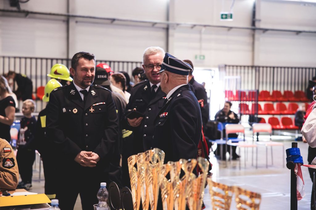 Trzech mężczyzn w mundurach strażackich rozmawia w hali z szarymi ścianami, z przodu złote puchary, w tle czerwone krzesła i ludzie.