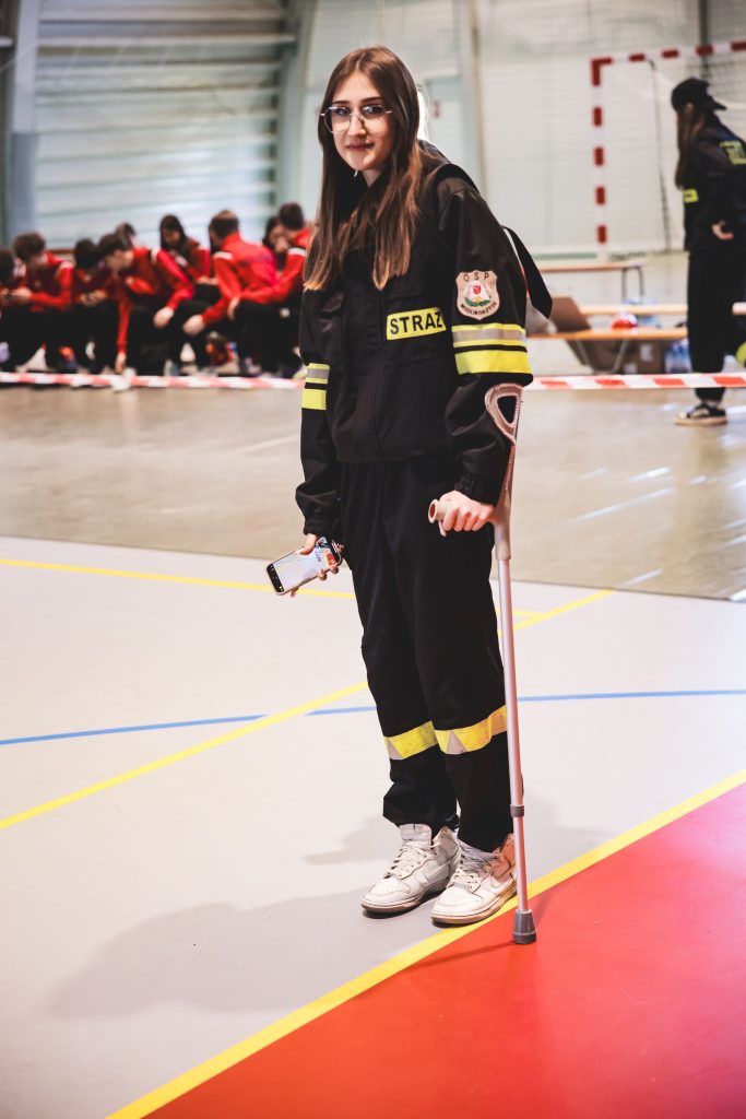 Młoda kobieta w czarnym mundurze strażackim z żółtymi odblaskami, okulary, biała laska podpierająca się, w ręku trzyma telefon, stoi na hali sportowej.