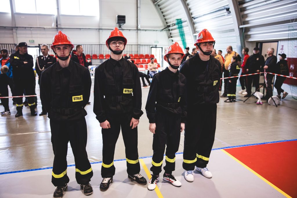 Czterech młodych strażaków w czarnych mundurach z żółtymi pasami i czerwonych hełmach stoi w rzędzie na hali sportowej, w tle ludzie i czerwone krzesła.