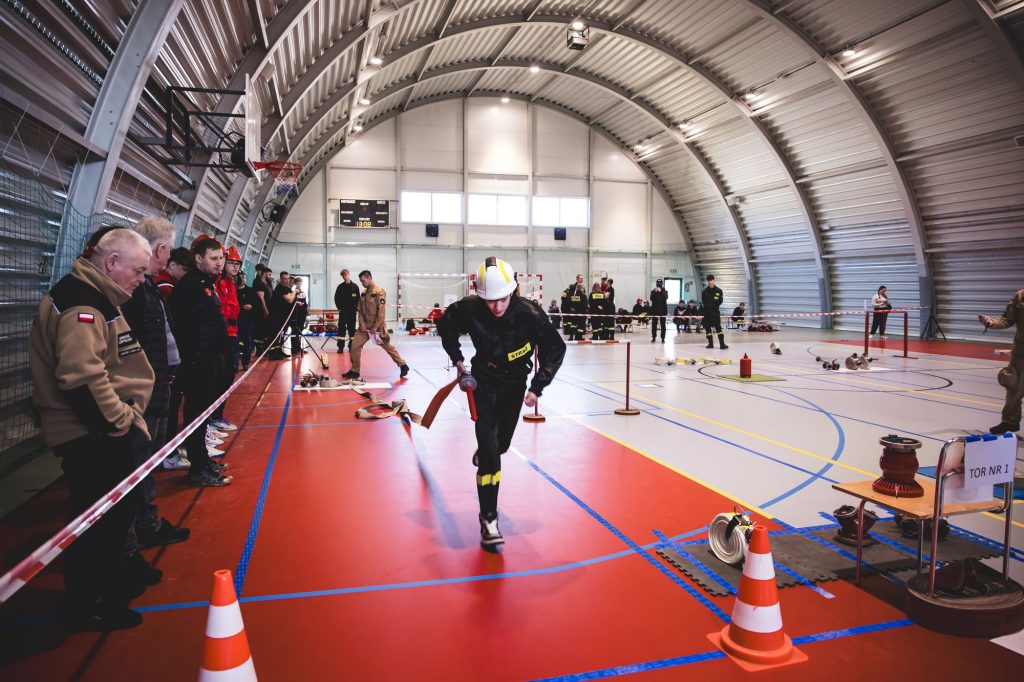 Mężczyzna w czarnym ubraniu strażackim i białym hełmie biegnie po czerwonej podłodze hali sportowej, trzymając składany wąż strażacki, obok stoi grupa obserwatorów.