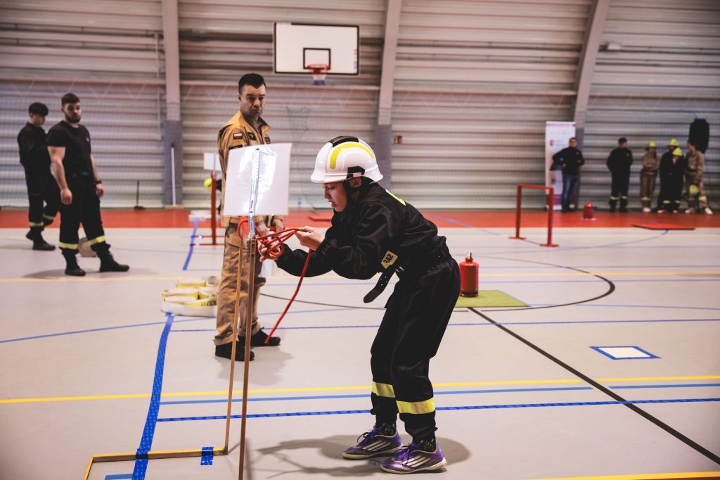 Osoba w czarnym ubraniu strażackim i białym kasku wiąże czerwony sznur na stojaku w hali sportowej, w tle inni ludzie w mundurach strażackich i kosz do koszykówki.