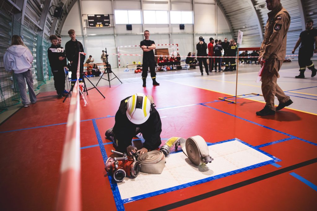 Osoba w hełmie i ubraniu strażackim na kolanach na czerwonej podłodze układa zrolowane węże strażackie, wokół inne osoby i sprzęt w hali sportowej.