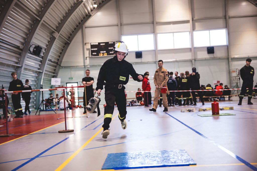 Młoda osoba w czarnym ubraniu strażackim i białym hełmie biegnie w hali sportowej, trzymając w ręku sprzęt pożarniczy, w tle ludzie i sprzęt na podłodze.