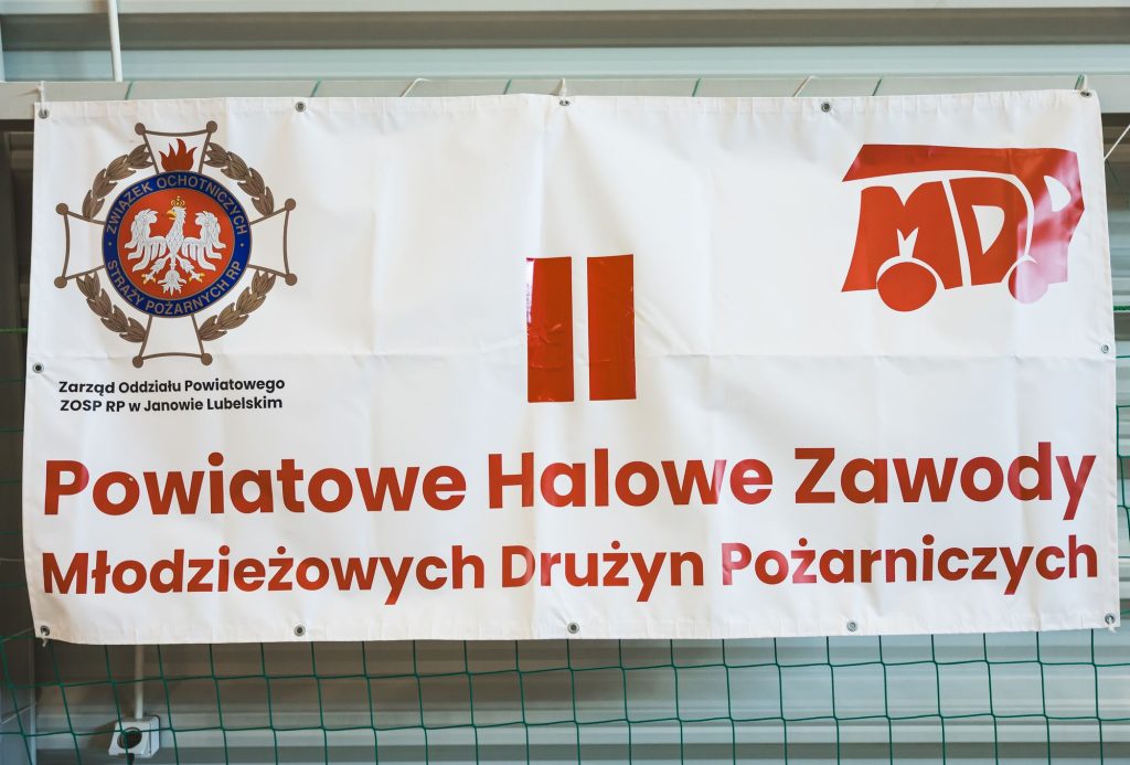 Biały baner z czerwonym i niebieskim logo, napisami w kolorze czerwonym i czarnym, na tle zielonej siatki i ściany z żaluzjami.