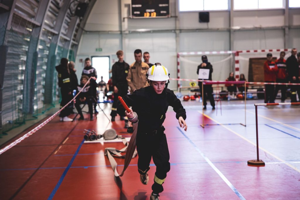 Chłopiec w czarnym ubraniu i białym kasku biegnie po sali sportowej z rozwiniętym wężem strażackim, w tle rozmyte postacie ludzi i sprzęt sportowy.