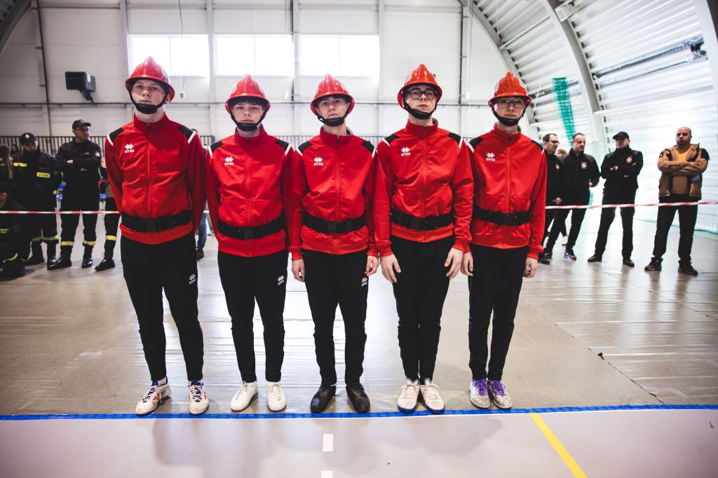 Pięciu młodych mężczyzn w czerwonych kaskach i kurtkach, czarnych spodniach i różnych butach stoi w rzędzie na hali sportowej, w tle kilka osób obserwujących.