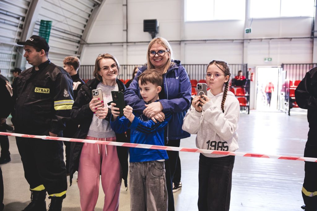 Grupa osób w hali sportowej, trzy kobiety i chłopiec z telefonami, dwaj mężczyźni w mundurach strażackich po bokach, czerwone krzesła i taśma zabezpieczająca.