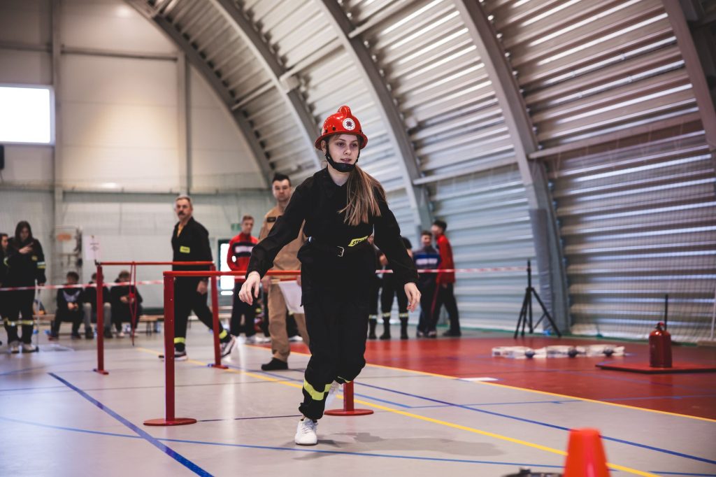Młoda kobieta w czarnym stroju strażaka i czerwonym hełmie biegnie na hali sportowej z przeszkodami, w tle ludzie w różnym ubraniu obserwują zawody.