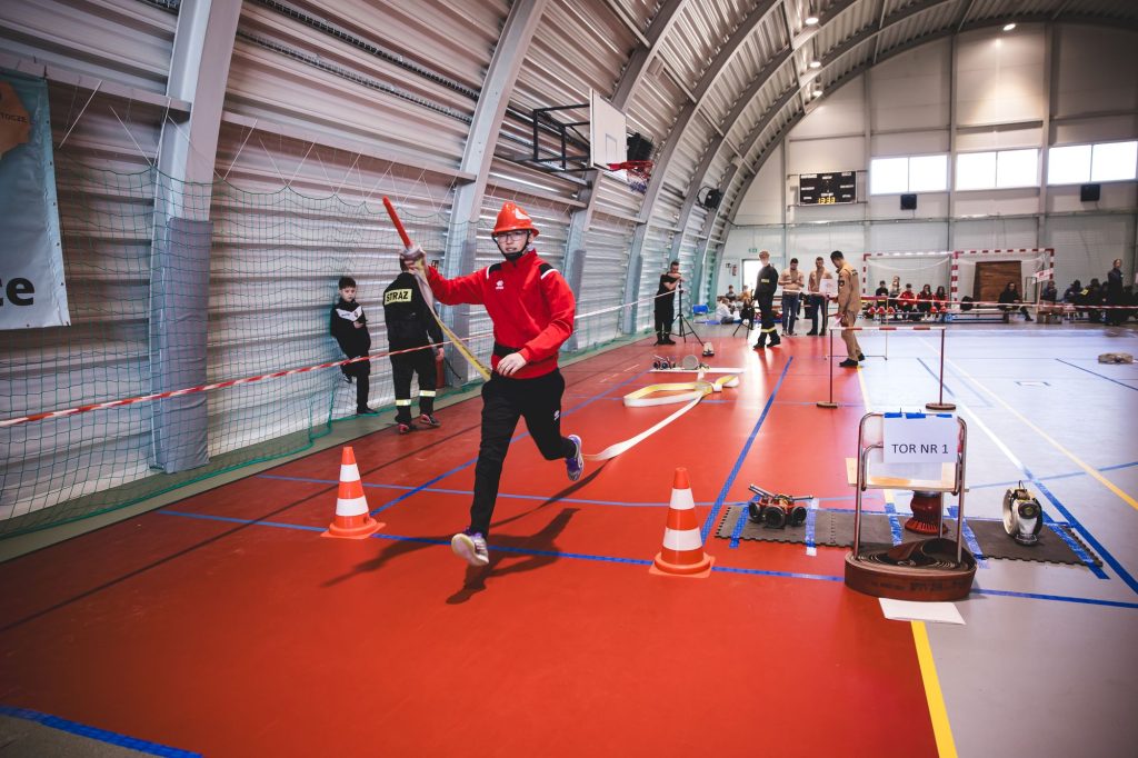 Chłopiec w czerwonej bluzie i pomarańczowym hełmie biegnie po czerwonej hali sportowej, trzymając zwinięty wąż strażacki, wokół stożki i sprzęt strażacki, w tle ludzie i siatka.
