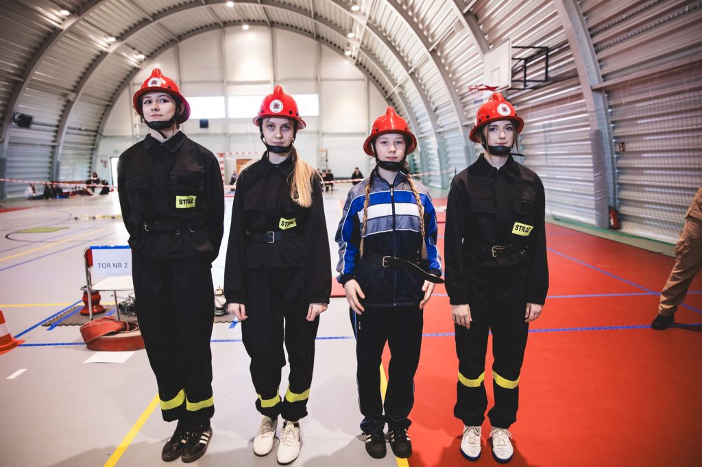 Cztery młode osoby w czerwonych hełmach, trzy w czarnych mundurach z żółtymi napisami „STRAŻ”, jedna w niebiesko-szarym stroju sportowym, stojące w hali sportowej.