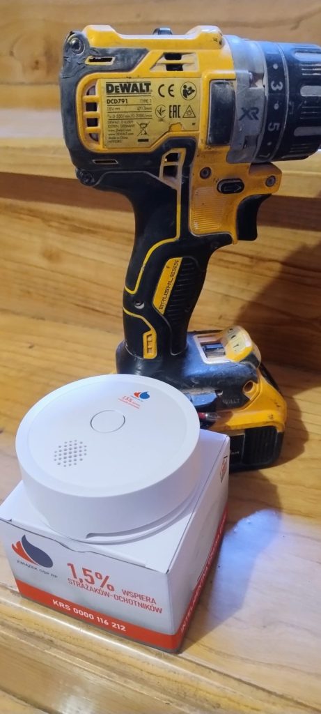 Żółto-czarna wiertarko-wkrętarka DeWalt stojąca na drewnianej powierzchni obok białego czujnika dymu na pudełku z napisem 1,5% wspiera strażaków-ochotników.