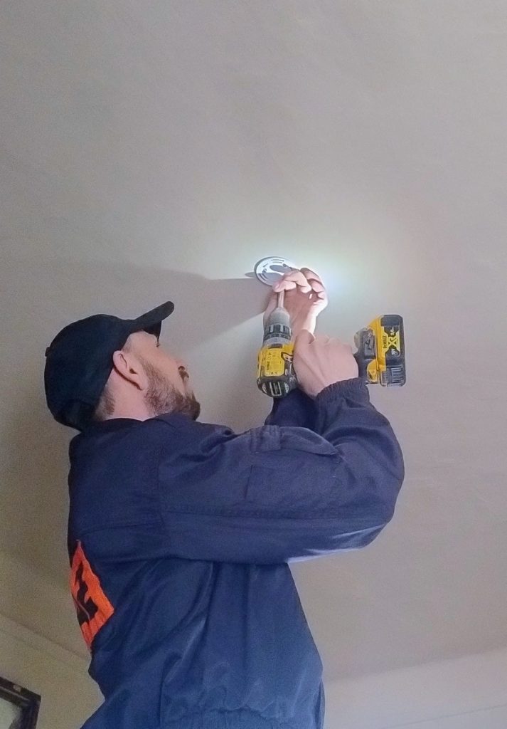 Mężczyzna w granatowej bluzie i czapce montuje biały czujnik dymu na jasnym suficie, używając żółtej wkrętarki.