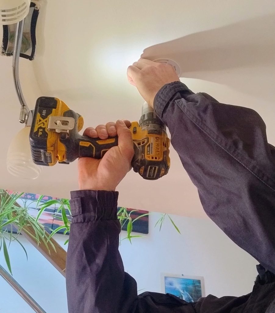 Ręce trzymające żółto-czarną wkrętarkę przy białym suficie, zielone liście roślin i obrazy na ścianie w tle.