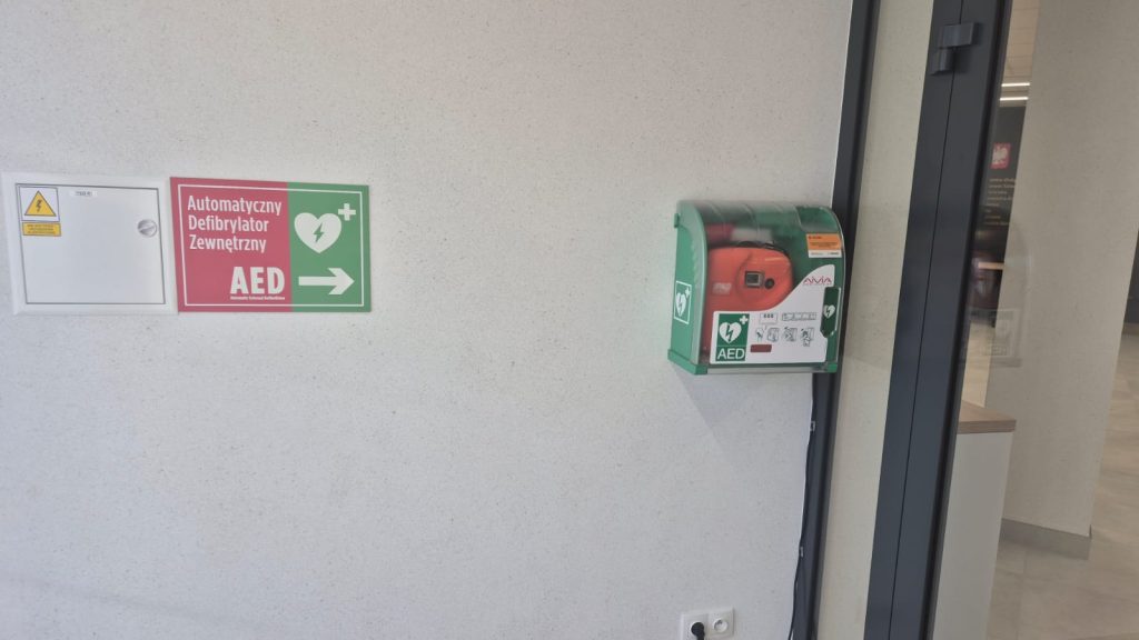 Zielona obudowa AED z pomarańczowym automatycznym defibrylatorem wewnątrz, biało-czerwono-zielona tablica informacyjna, szara ściana, fragment szarej ramy drzwi.