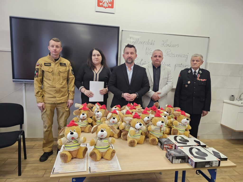 Pięć osób stoi za stołem z pluszowymi maskotkami w kaskach strażackich, certyfikatami i opakowaniami słuchawek, tablica z napisem konkurs, biało-czerwony orzeł.