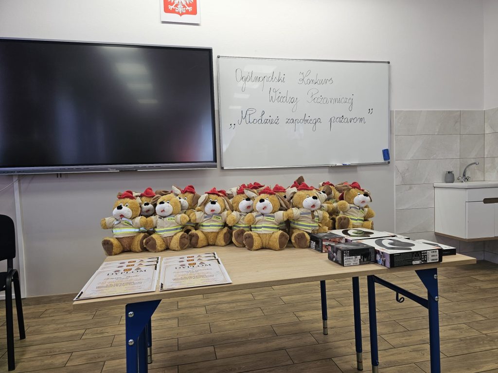 Pluszowe misie w żółtych strażackich kurtkach z czerwonymi czapkami, dyplomy i nagrody na drewnianym stole, za nim ekran i tablica z napisem konkursowym, jasne ściany.