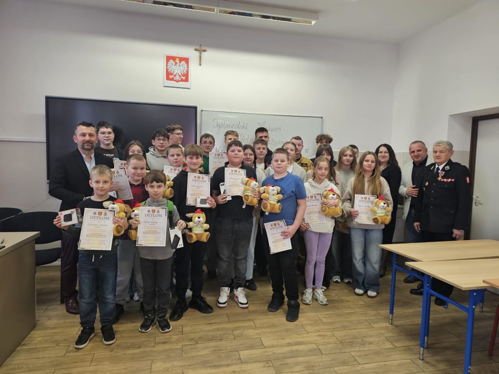 Grupa dzieci i dorosłych w sali z białą tablicą, dzieci trzymają dyplomy i pluszowe misie, na ścianie polski godło i krzyż, podłoga drewniana.