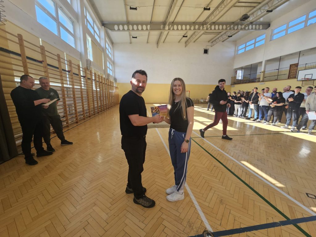 Dwóch uśmiechniętych ludzi w sportowym stroju podaje sobie ręce w jasnej sali gimnastycznej z drewnianą podłogą, w tle stoi grupa osób obserwujących wydarzenie.