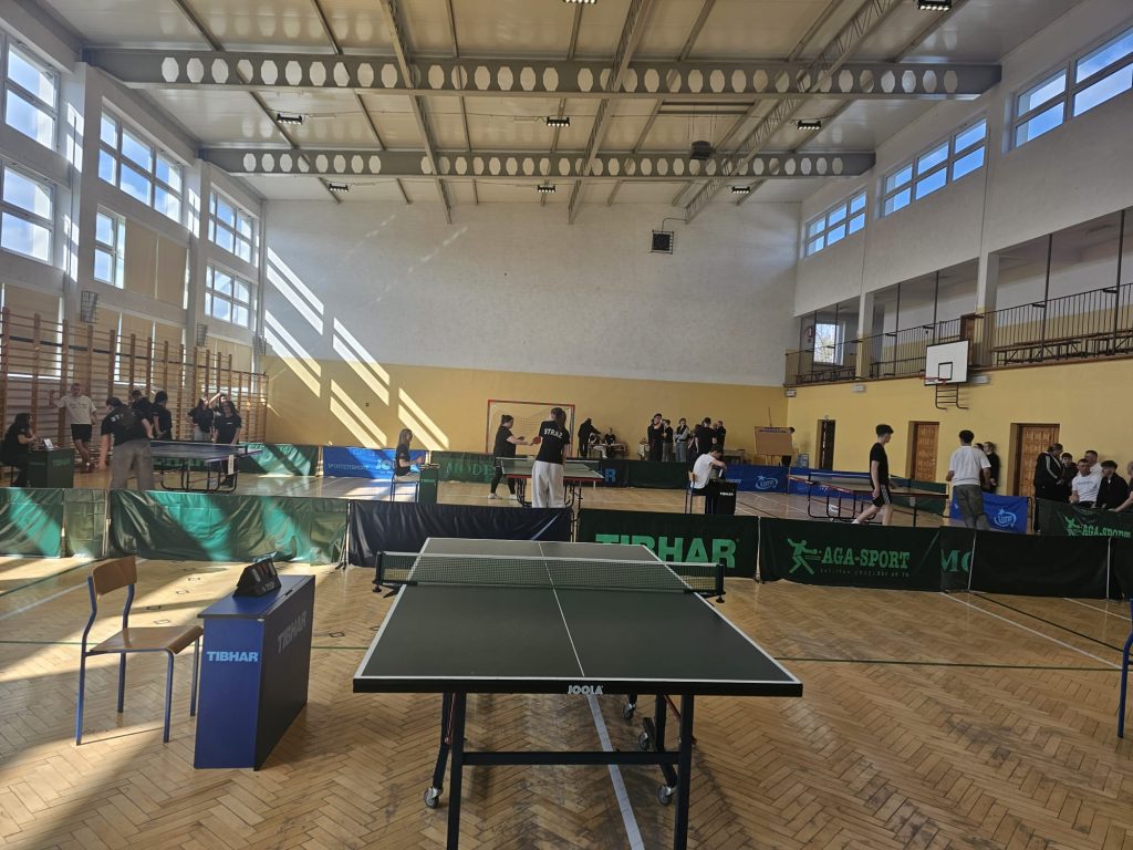 Hala sportowa z dużymi oknami, kilkoma stołami do tenisa stołowego, ludzie grający i siedzący, drewniana podłoga, jasne ściany, siatki i barierki reklamowe.