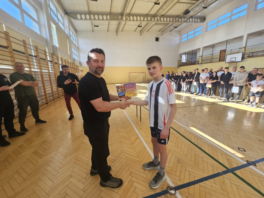 Dwóch mężczyzn ściska dłonie, jeden trzyma żółty dyplom, w tle grupa ludzi stojąca w sali gimnastycznej z dużymi oknami i drewnianą podłogą.