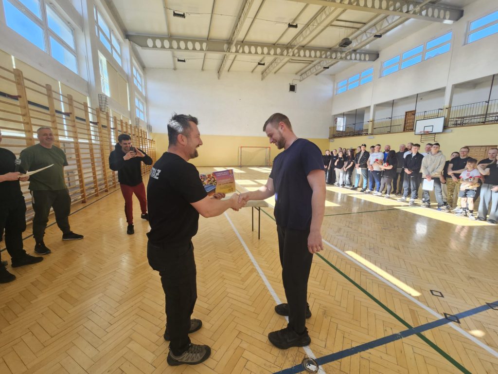 Dwóch mężczyzn uściski dłonie w hali sportowej, w tle grupa ludzi w rzędzie, drewniana podłoga, wysoka przestrzeń z dużymi oknami i sprzętem sportowym.