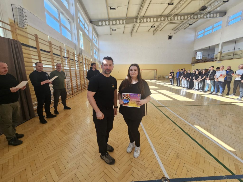 Grupa ludzi stojących w hali sportowej na drewnianej podłodze, dwie osoby na pierwszym planie trzymające kolorowy dyplom, jasne światło wpada przez okna.