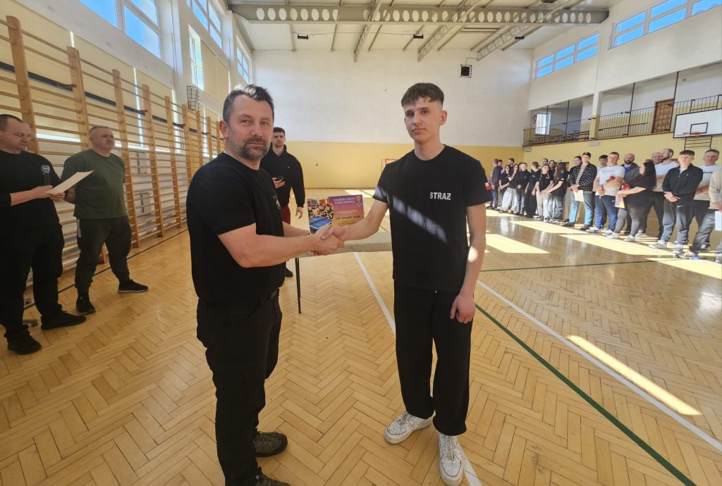 Dwóch mężczyzn w czarnych ubraniach ściska dłonie na drewnianej podłodze hali sportowej, w tle stoją ludzie i tablica z napisem.