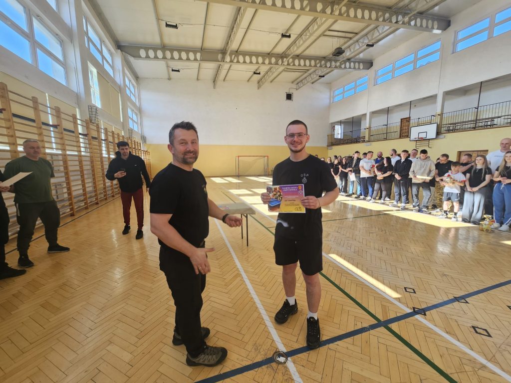 Dwóch mężczyzn w czarnych ubraniach na drewnianej sali gimnastycznej, jeden trzyma kolorowy dyplom, w tle grupa stojących ludzi, wysokie okna i siatka bramki.