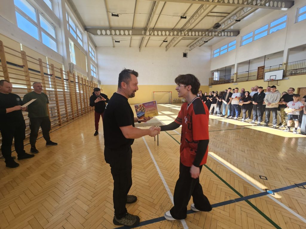 Mężczyzna w czarnym ubraniu wręcza dyplom chłopcu w czerwonej koszulce na drewnianej podłodze hali sportowej, w tle stoją ludzie obserwujący uroczystość.