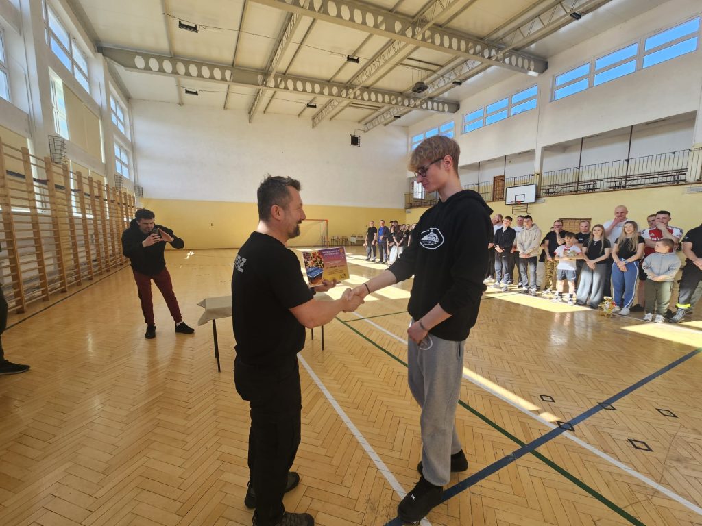 Dwóch mężczyzn na drewnianej podłodze hali sportowej podaje sobie ręce, w tle grupa osób stojąca w rzędzie, jeden mężczyzna robi zdjęcie.