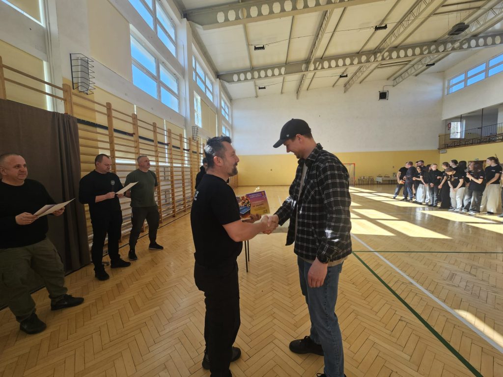 Dwóch mężczyzn ściska sobie dłonie, jeden trzyma kolorową kartkę, w tle grupa ludzi stojąca w hali sportowej z drewnianą podłogą i dużymi oknami.