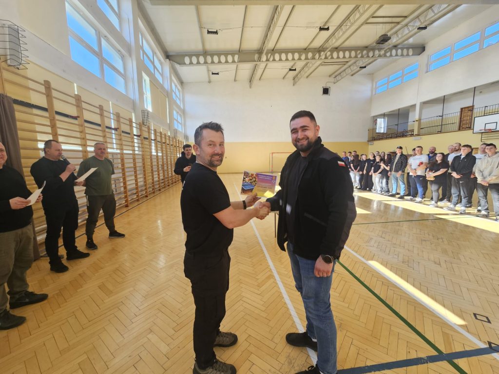 Dwaj mężczyźni uściskiem dłoni, jeden trzyma kolorowy dyplom, w hali sportowej z drewnianą podłogą, w tle stoją ludzie i mężczyźni z dokumentami.