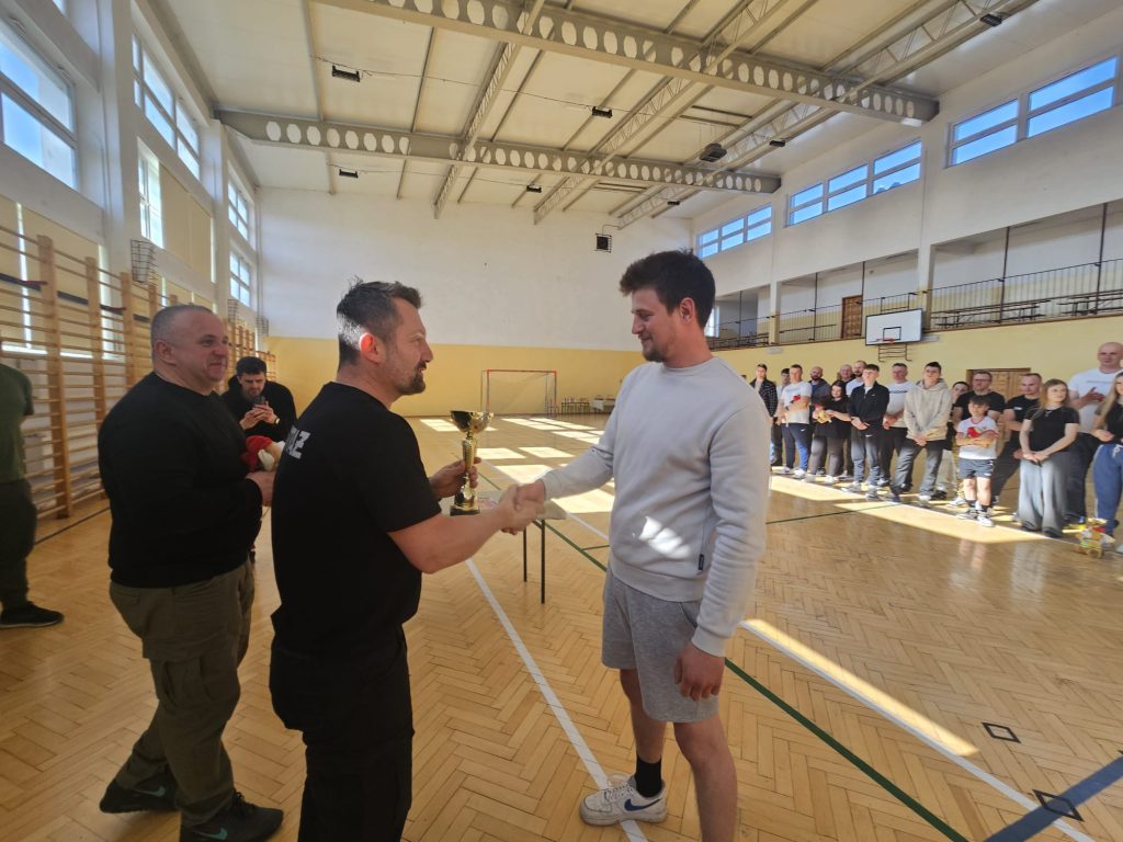 Dwóch mężczyzn ściska się dłonią w hali sportowej, jeden trzyma złoty puchar, w tle grupa ludzi stoi przy ścianie.