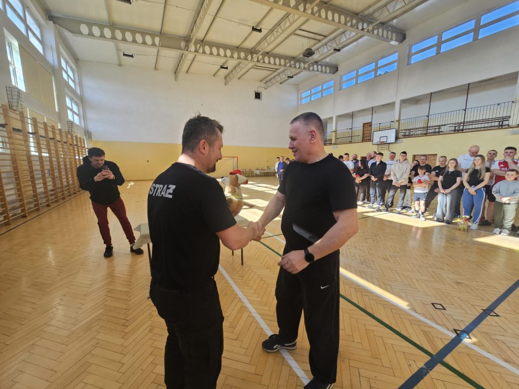 Dwóch mężczyzn ściska dłonie w hali sportowej, jeden w czarnej koszulce z napisem STRAŻ, w tle grupa osób ustawionych w rzędzie, parkiet, wysokie okna i sprzęt sportowy.